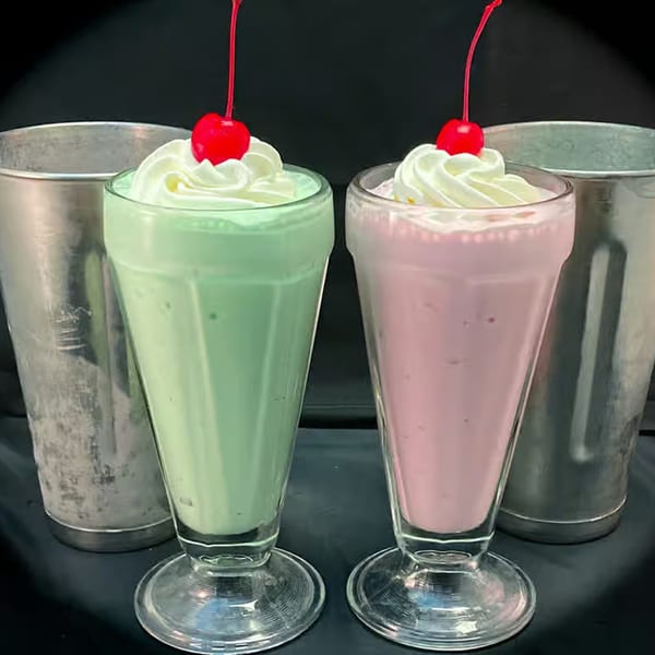 Two milkshakes, one green and one pink, topped with whipped cream and cherries, with metal shakers.