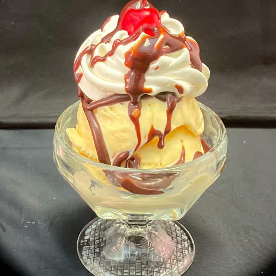 Vanilla ice cream sundae with whipped cream, chocolate syrup, and a cherry in a glass dish.